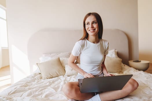 A woman with long hair smiles while seated cross-legged on a bed. She uses her laptop in a well-lit bedroom filled with soft furnishings. Natural light creates a warm atmosphere.