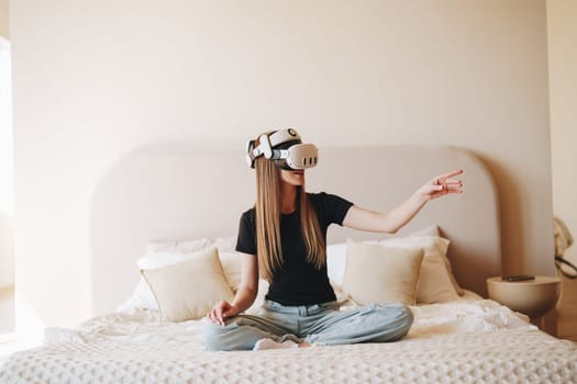 A young woman is seated cross-legged on a bed, wearing a virtual reality headset and fully engaged in the immersive experience.