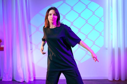 A dancer showcases her movement in a contemporary studio, illuminated by vibrant colored lights creating an artistic ambiance. The relaxed outfit enhances her fluidity.