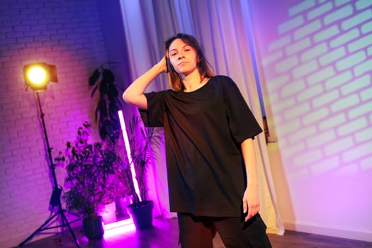 A person stands dramatically in a colorful indoor space, illuminated by purple and yellow lights. Stylish plants surround her as she strikes a confident pose, creating an artistic atmosphere.