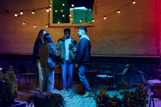 A group of four friends socialize outdoors during the evening, surrounded by string lights. They share drinks and laughter in a cozy, casual ambiance.