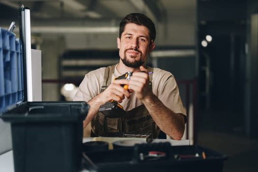 A focused worker uses a power tool while seated at a cluttered bench in a well-lit workshop. Various tools and storage bins are nearby, suggesting an active work environment.