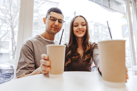 A young couple smiles while sitting at a cafe table, holding disposable cups filled with drinks. The backdrop shows a busy city street through the window.