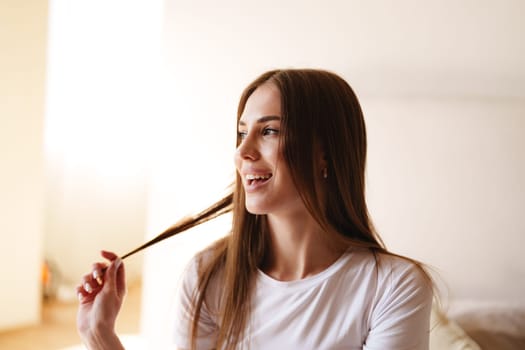A young woman laughs while playing with her long hair, sitting in a well-lit room. Sunlight streams through the window, creating a cheerful atmosphere.