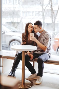 Two individuals share a tender moment in a cafe, sipping warm beverages while snow gently falls outside. They sit closely, creating a warm and intimate atmosphere.