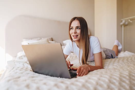 A young woman is comfortably lying on her bed, focused on her laptop. The setting is a bright bedroom with soft lighting. She appears engaged and content while working.