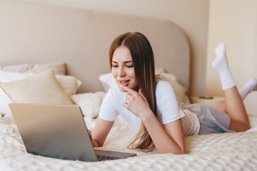 A woman with long hair lies comfortably on her stomach on a bed, engaged in an online activity on her laptop. Natural light illuminates the serene space, creating a peaceful atmosphere.