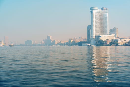 The calm waters reflect the tall buildings of Cairo under a soft morning light. The skyline features modern architecture and hints of bustling city life beginning to awaken.