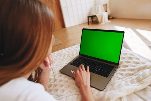 A woman sits on a bed covered with a soft blanket, intently working on a laptop displaying a bright green screen. Sunlight streams through the room, creating a warm atmosphere.