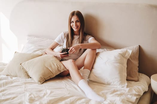 A woman sits on her bed with plush pillows, casually holding her phone and smiling. Natural light floods the room, creating a warm and inviting atmosphere.