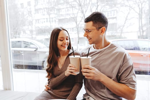 A couple sits closely by a large window in a cafe, smiling and holding warm drinks. Outside, snow is visible, enhancing the cozy atmosphere of their day together.