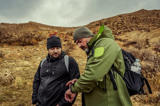Two men dressed in outdoor gear stand on a trail discussing their navigation tools while surrounded by a rugged, mountainous terrain under a gloomy sky.