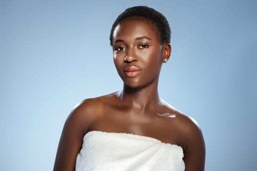A woman with short hair stands confidently, wearing a white towel. She is against a soft blue background that enhances her natural beauty and calm demeanor. The lighting is warm and flattering.