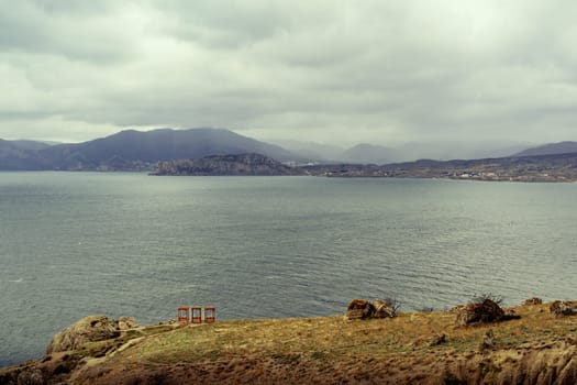 Calm waters stretch across the landscape beneath a cloudy sky, framed by rugged mountains and a rocky shoreline. The atmosphere is serene, evoking a sense of peace and solitude.