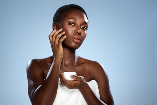 A woman stands in a well-lit studio, gently applying a skincare product on her cheek while holding a small container. Her smooth skin glows in the soft light.