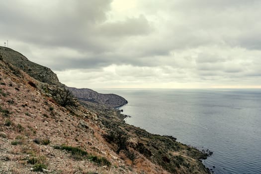 Lush cliffs descend into a calm sea on a cloudy day, showcasing the natural beauty of the coastal region. The landscape reveals a serene atmosphere and earthy tones.