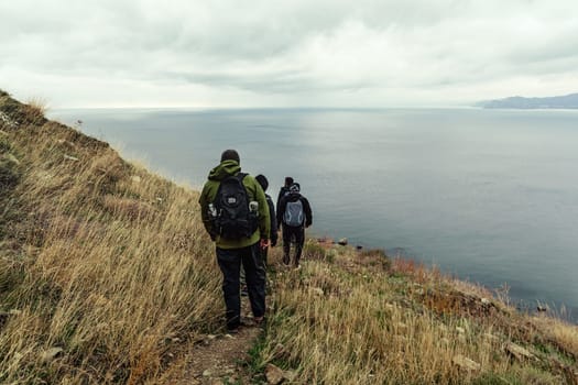 Two hikers navigate a rugged coastal trail surrounded by tall grasses, as gray clouds linger overhead and the ocean stretches beyond the path.