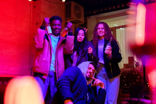 Four friends gather in a lively indoor space, illuminated by colorful lights, holding drinks and enjoying a joyful celebration together in the evening.