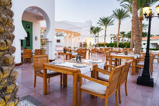 This setting features elegantly arranged wooden tables and chairs under a tranquil sky. Lush palm trees surround the outdoor dining space, creating a serene atmosphere.
