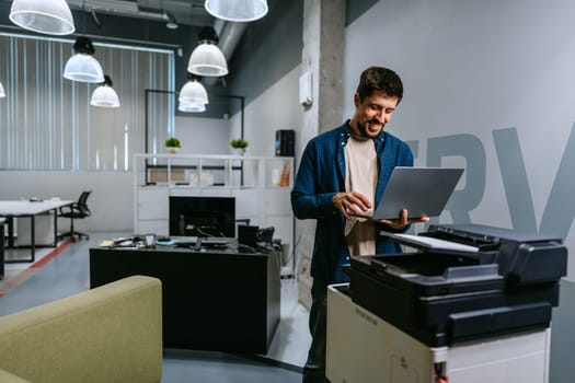 A man stands in a contemporary office setting, interacting with a laptop while positioned next to a printer. The area features a vibrant atmosphere with various workspaces.