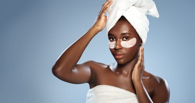 A young woman with a towel wrapped around her hair applies an under-eye mask in a simple studio. The soft blue background enhances her natural beauty.
