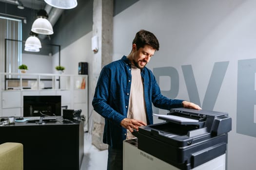 A man in a blue checkered shirt uses a printer in a contemporary office space filled with natural light. The atmosphere is relaxed, emphasizing productivity and collaboration.