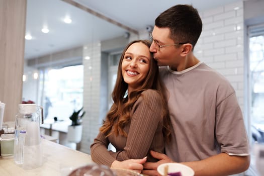 A couple enjoys an intimate moment in a charming cafe. The man leans in to kiss the woman on the cheek, who beams with happiness. Soft natural light pours in through the windows.