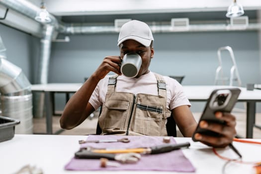 A focused worker in casual attire sits at a table in a contemporary workspace, sipping coffee from a mug and using a smartphone. Tools and materials are neatly arranged.