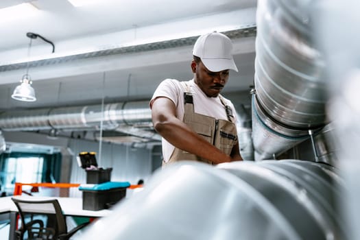 A worker in beige overalls and a white cap is focused on repairing equipment in a well-lit workshop. The environment features metal ducting and workstations.