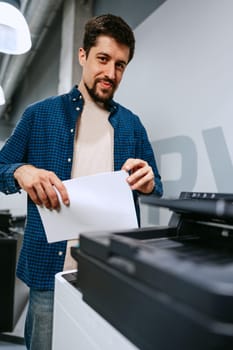 A man in a blue checkered shirt and beige t-shirt stands in a contemporary office, holding a sheet of paper as he interacts with a printer. The setting is bright and spacious.