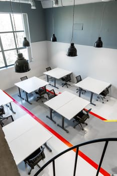 A spacious office interior showcases multiple white tables and ergonomic chairs arranged in a functional layout. The bright space is illuminated by large windows and hanging lights.