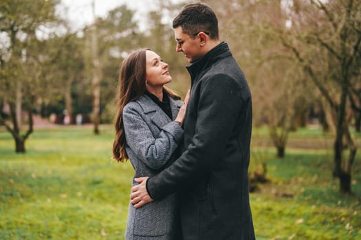 A couple stands close together in a green park, gazing into each other's eyes. They share a warm connection, surrounded by nature's beauty on an overcast day.