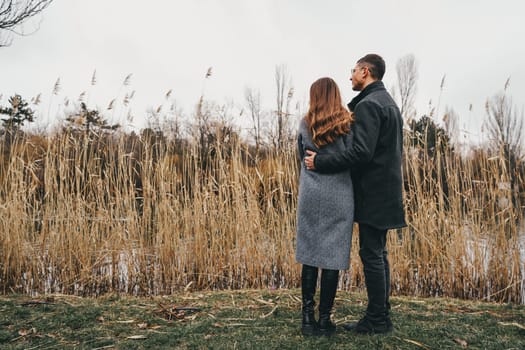 Two individuals stand close together, embracing gently as they gaze towards a serene landscape. Tall grasses sway in the wind near a calm body of water, creating a peaceful atmosphere.