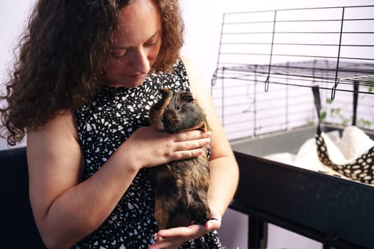 A woman gently holds a guinea pig in her arms while sitting in a comfortable indoor environment. A pet cage is visible in the background, indicating a warm atmosphere.