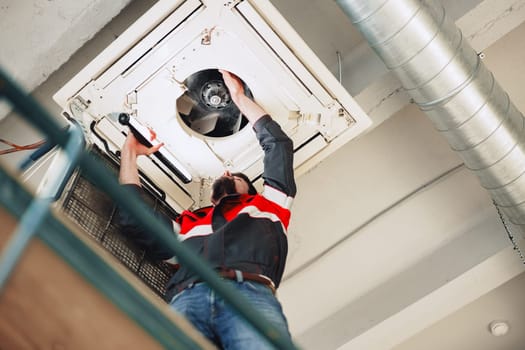A technician is reaching up to repair a ventilation system in a commercial building. The setting features exposed ductwork and an industrial aesthetic, illuminated by natural light.