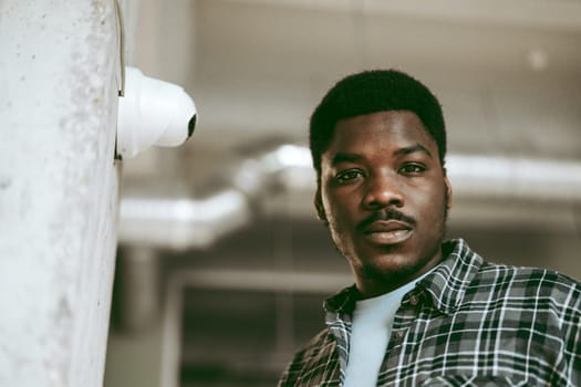 A young man stands near a wall with a surveillance camera in an industrial-style location. He embodies confidence with a thoughtful expression, surrounded by soft, natural light.