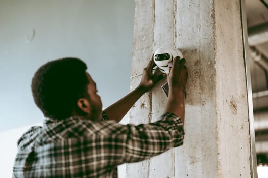 A technician focuses on installing a white security camera on a concrete pillar inside a bright, modern office. Tools and equipment are nearby as he works diligently.