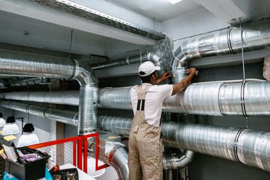 A worker is focused on installing ventilation ductwork in an industrial space. The surroundings feature silver pipes and construction elements, indicating an active project.
