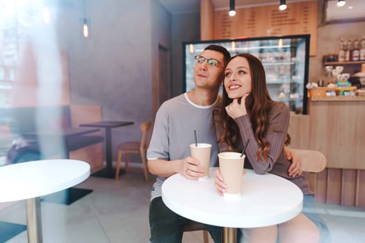 A young couple sits at a small table in a bright cafe, holding cups of coffee and smiling. They share a moment of connection while enjoying their time together.