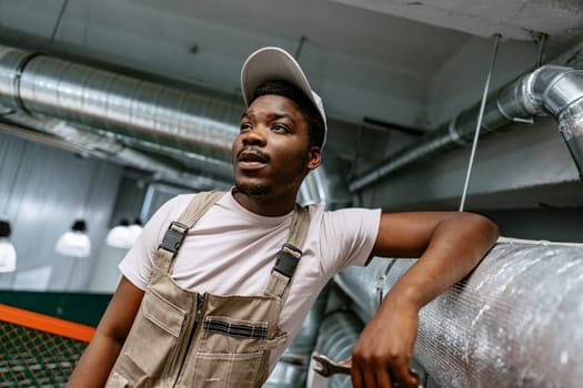 A young man in beige overalls and a cap leans against a metal railing in a modern industrial space, showcasing a relaxed demeanor and style.
