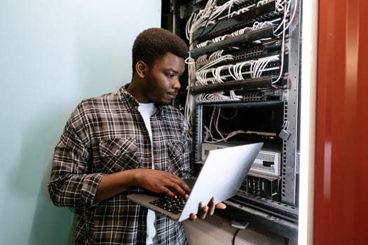 A technician is focused on adjusting server settings while holding a laptop in a data center. Cables and servers create a busy yet organized atmosphere in the background.