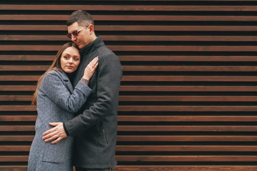 A couple stands close, sharing a warm embrace against a backdrop of wooden slats. Their clothing suggests a cool day, and their expressions reflect comfort and affection for one another.