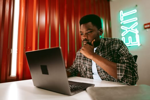 A young man sits at a desk, deeply engaged with his laptop in a stylish workspace. Warm lighting illuminates the environment, enhancing concentration.