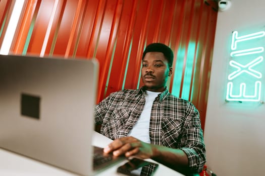 A young man sits focused in a trendy workspace, engaging with his laptop. The vibrant red wall and neon exit sign create a contemporary atmosphere during the evening.