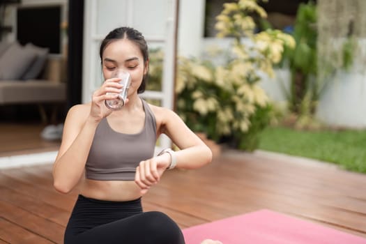 A young woman sits in a yoga pose, drinking water and checking her watch, promoting wellness and mindfulness in her daily routine.