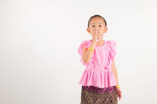Portrait Thailand kid girl smiling traditional Thai dress costume making finger on lips mouth silent quiet gesture, studio isolated white background, kindergarten child marking silence for stop quiet
