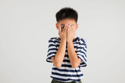 Asian portrait young kid boy in depressed bad mood covering face with hands and peering out with one eye between fingers studio shot isolated on white background, primary child surprised and shocked