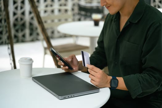 Man holding credit card and using smartphone for an online transaction at an outdoor cafe.