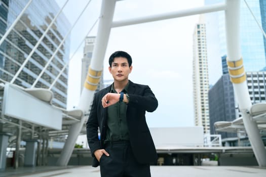 Confident businessman in a black suit looking at his smartwatch while standing in a modern urban.