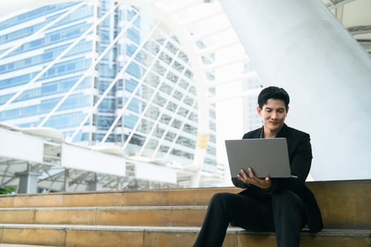 Businessman in formal attire uses laptop while sitting on city steps surrounded by skyscrapers.